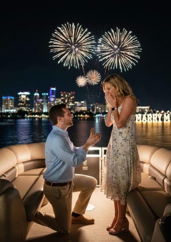 A man kneels to propose to a surprised woman on a boat at night, with fireworks in the sky and city lights in the background. The words MARRY ME? are illuminated on the shoreline behind them.