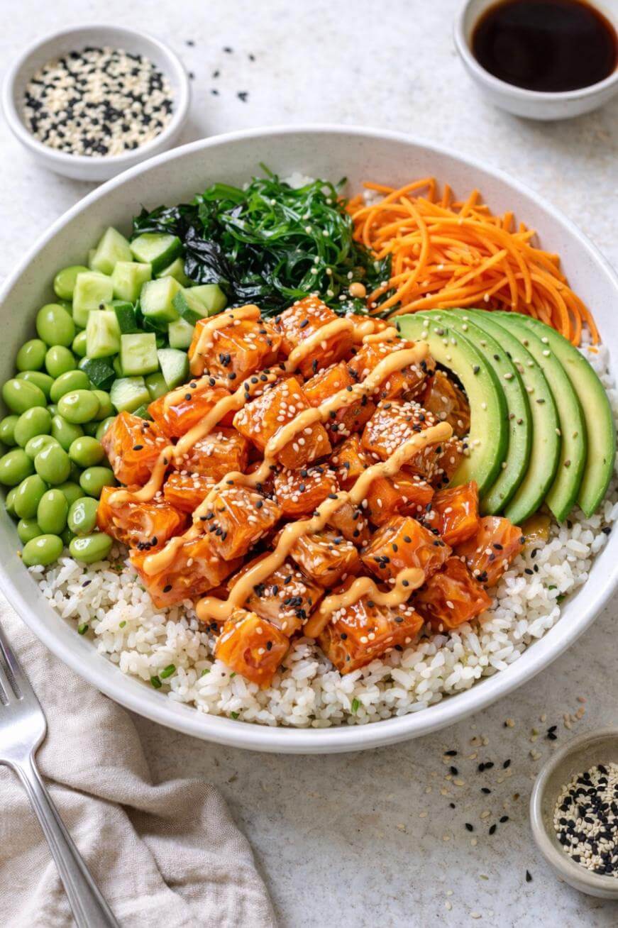 A poke bowl with diced salmon in sauce, white rice, sliced avocado, shredded carrots, chopped cucumber, edamame, seaweed salad, and sesame seeds, drizzled with creamy dressing, served with a fork and small bowls of soy sauce and sesame seeds.