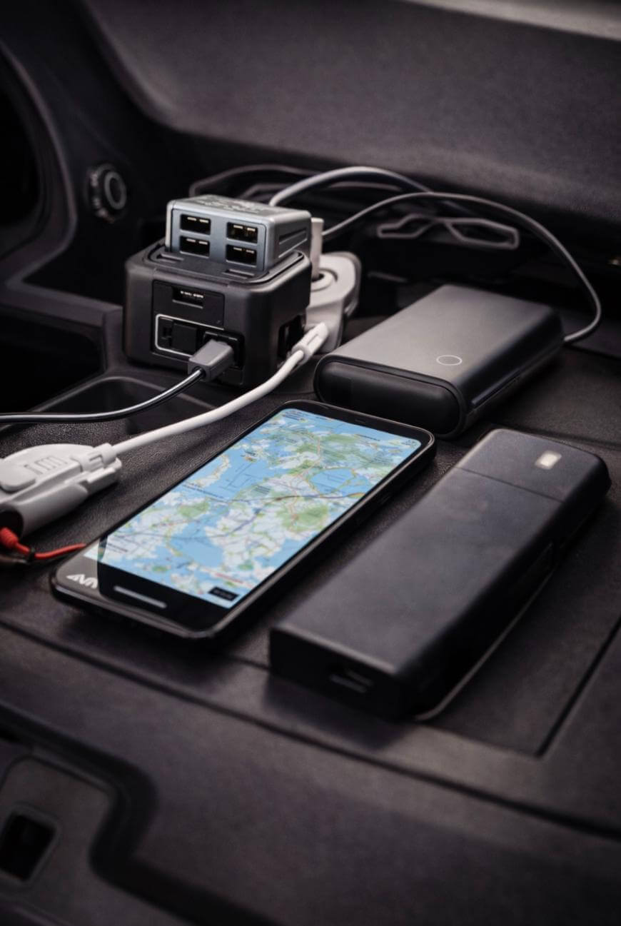 A smartphone displaying a map, two black power banks, and a multi-port USB charger with several cables are placed on a car dashboard.
