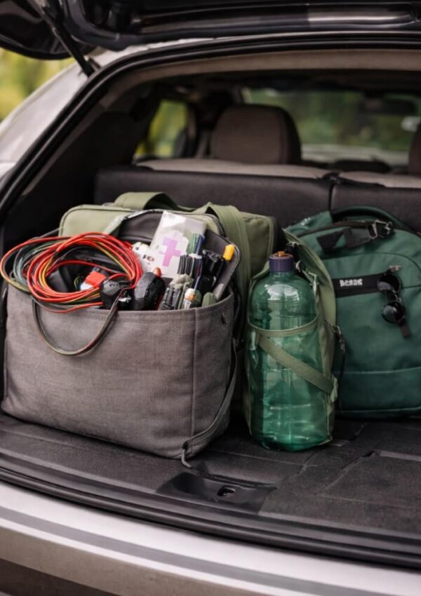 An open car trunk showing a gray bag with jumper cables, a first aid kit, and tools, a large green water bottle, and a green backpack with sunglasses attached.