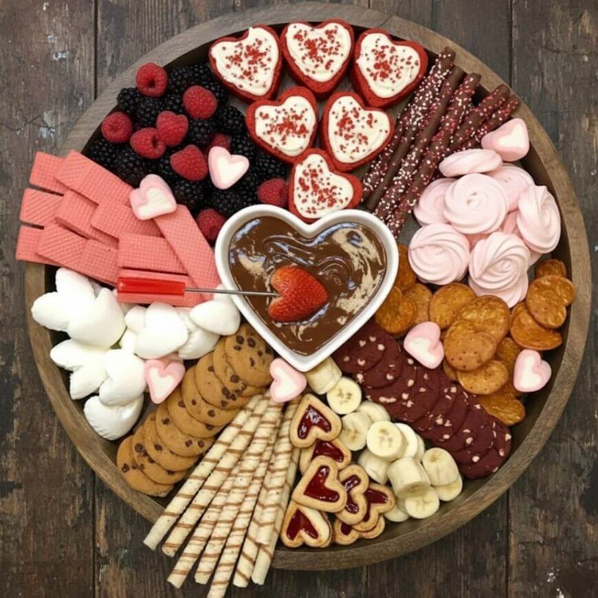 A round wooden platter filled with assorted sweets, heart-shaped cookies, wafers, berries, marshmallows, pretzels, banana slices, and a heart-shaped bowl of chocolate dip with a strawberry.