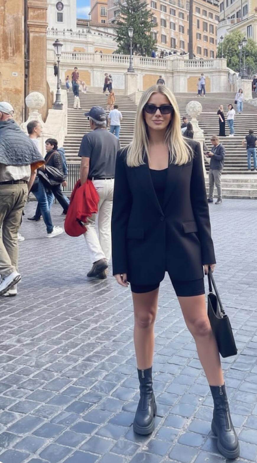 A woman wearing a black blazer, black shorts, and black boots stands on a cobblestone street in front of large stone steps, surrounded by people and historic buildings during the day.