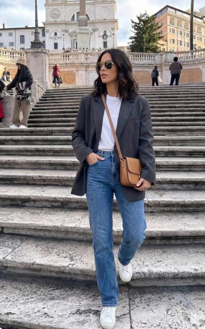 A woman in sunglasses, a gray blazer, white t-shirt, blue jeans, and white sneakers stands on stone steps outdoors with a tan crossbody bag. Historic buildings and other people appear in the background.