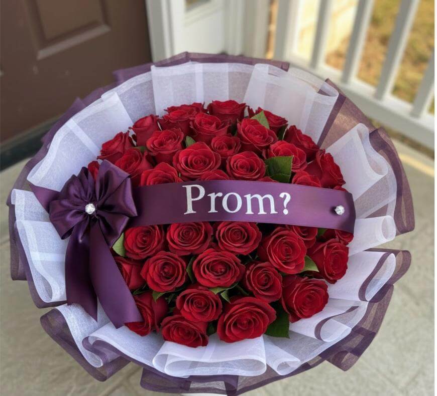 A bouquet of red roses wrapped in white and purple tulle with a purple ribbon that says Prom? in white letters, and a matching purple bow on the side.