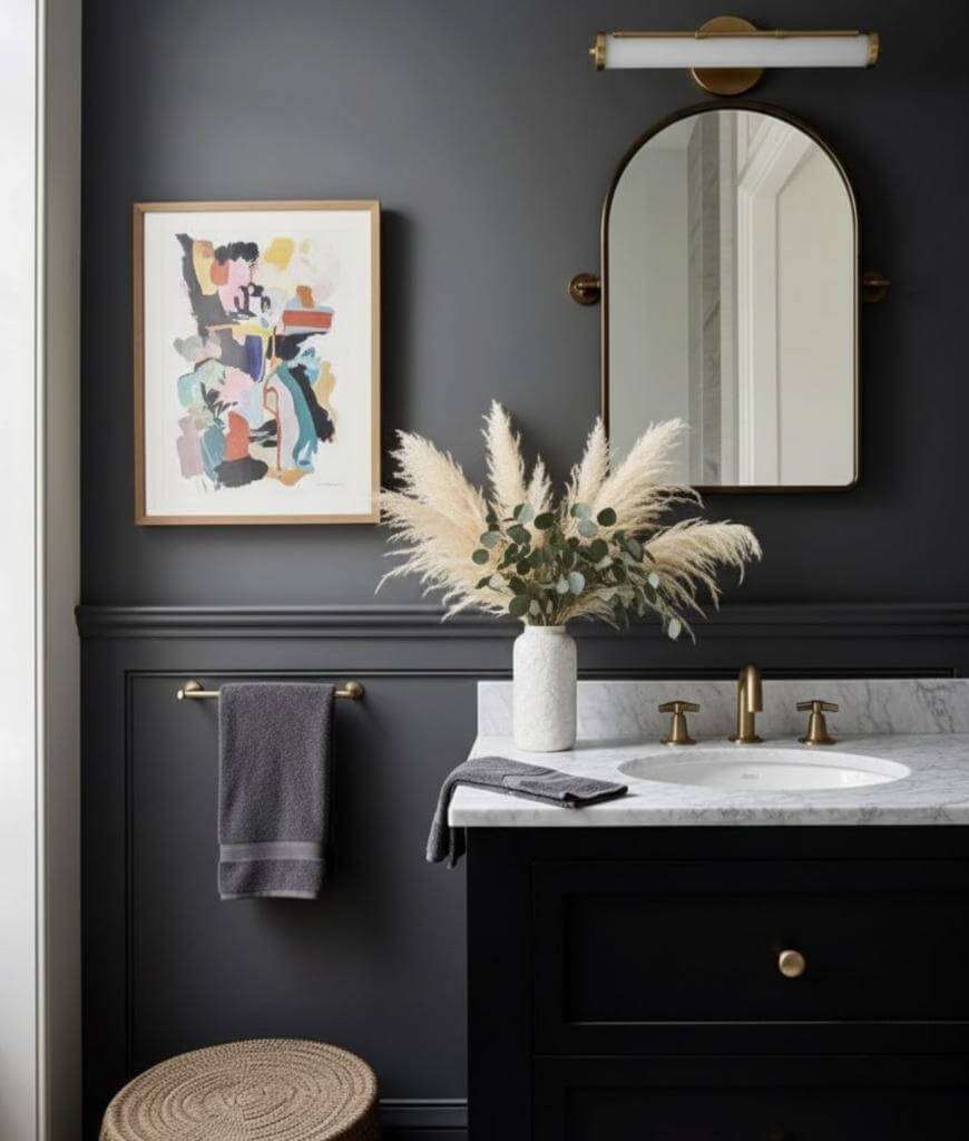 A modern bathroom with dark walls, a round mirror, brass fixtures, a white marble countertop, a vase of pampas grass, a hand towel, a woven stool, and abstract artwork on the wall.
