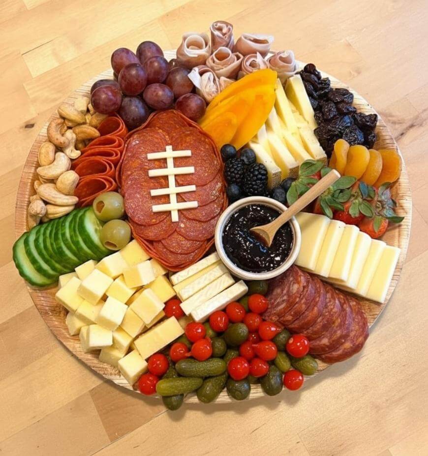 A round charcuterie board with grapes, cheese, meats, cucumbers, strawberries, cherry tomatoes, pickles, dried fruits, nuts, olives, and a small bowl of jam. Slices of meat are arranged like a football.