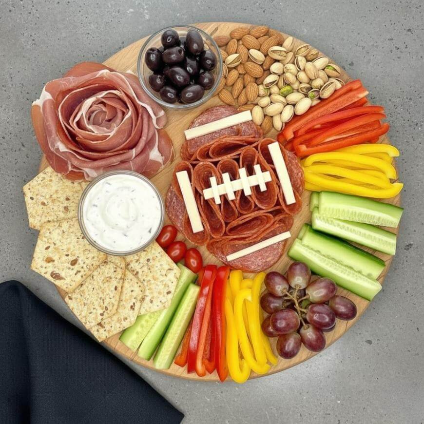 A round wooden platter with meats, cheese, crackers, dip, olives, nuts, sliced bell peppers, cucumber sticks, grapes, and dip. Salami and cheese are arranged like a football in the center.