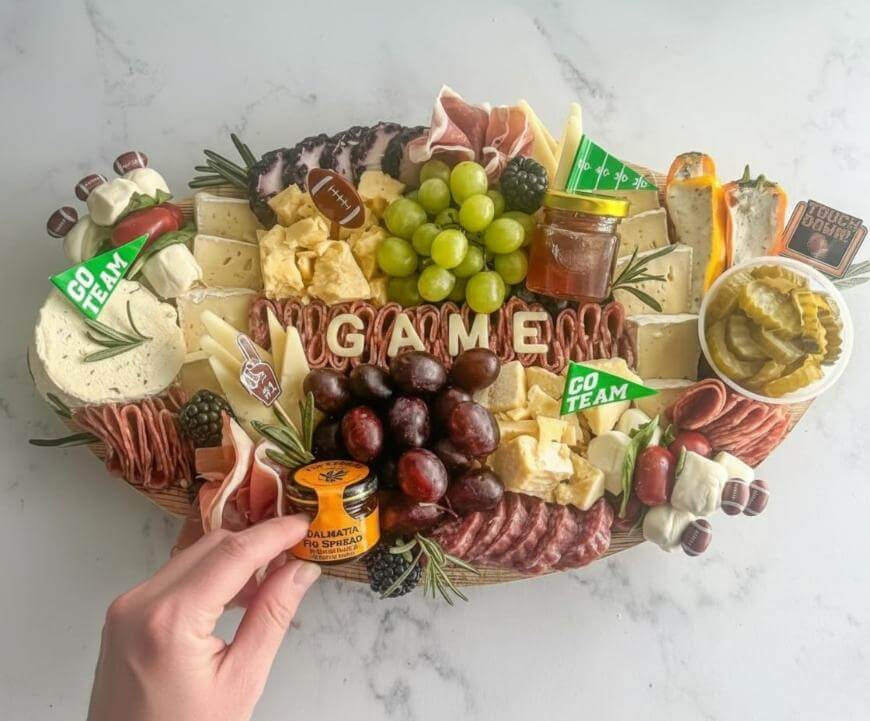 A football-themed charcuterie board with meats, cheeses, crackers, grapes, pickles, fruit, and dips. The word GAME is spelled out in cheese; food picks say GO TEAM. A hand holds a small container of Dalmatia fig spread.