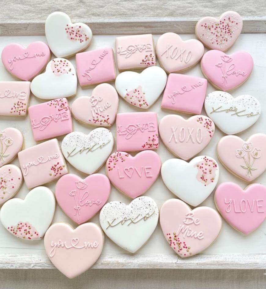 Assorted heart and rectangle sugar cookies decorated with pink and white icing, featuring messages like Love, XOXO, Be Mine, and you + me, as well as floral designs and sprinkles.