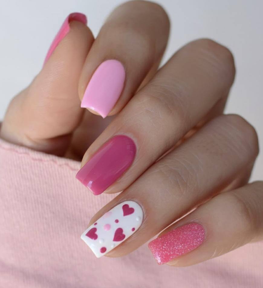 A hand with manicured nails painted in different shades of pink. One nail features white polish with small pink and red heart designs, while another has a sparkly pink finish. The person wears a light pink sleeve.