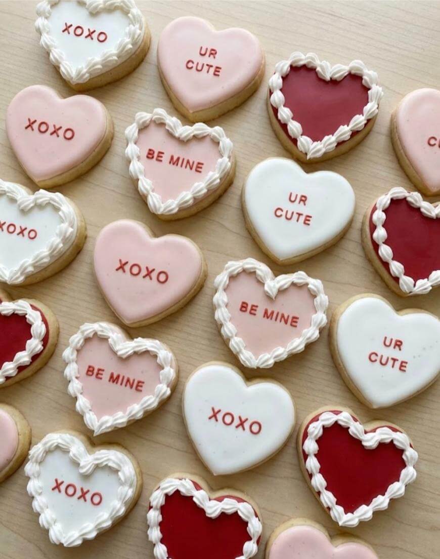 Heart-shaped cookies decorated in red, pink, and white icing, with messages like “XOXO,” “BE MINE,” and “UR CUTE” written on them, arranged on a light wooden surface.