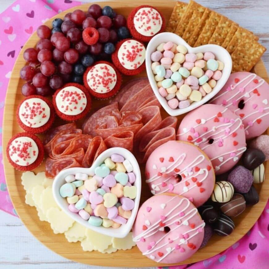 A wooden platter holds pink donuts, candy hearts in heart-shaped bowls, red grapes, mini cupcakes with sprinkles, crackers, chocolates, and sliced cured meat, arranged for a festive treat.