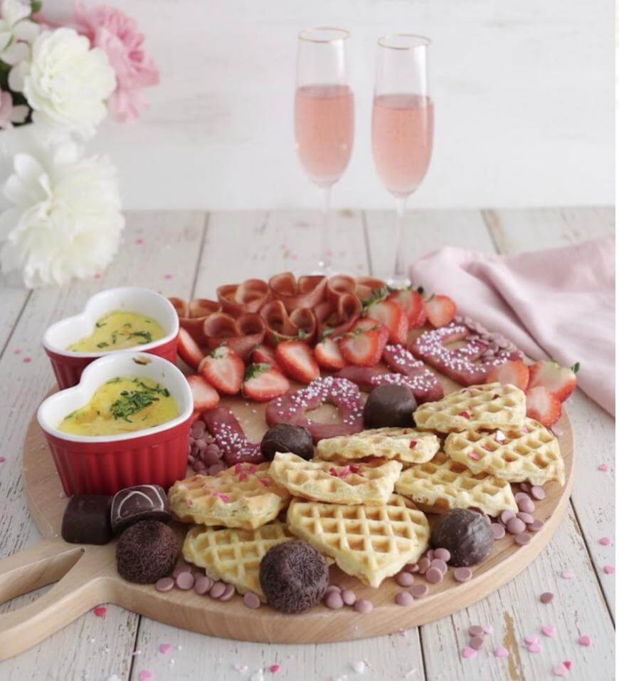 A wooden board with heart-shaped waffles, chocolate truffles, strawberries, rolled ham, two ramekins of baked eggs, pink candy sprinkles, and two glasses of pink sparkling drink, set on a light table with flowers.