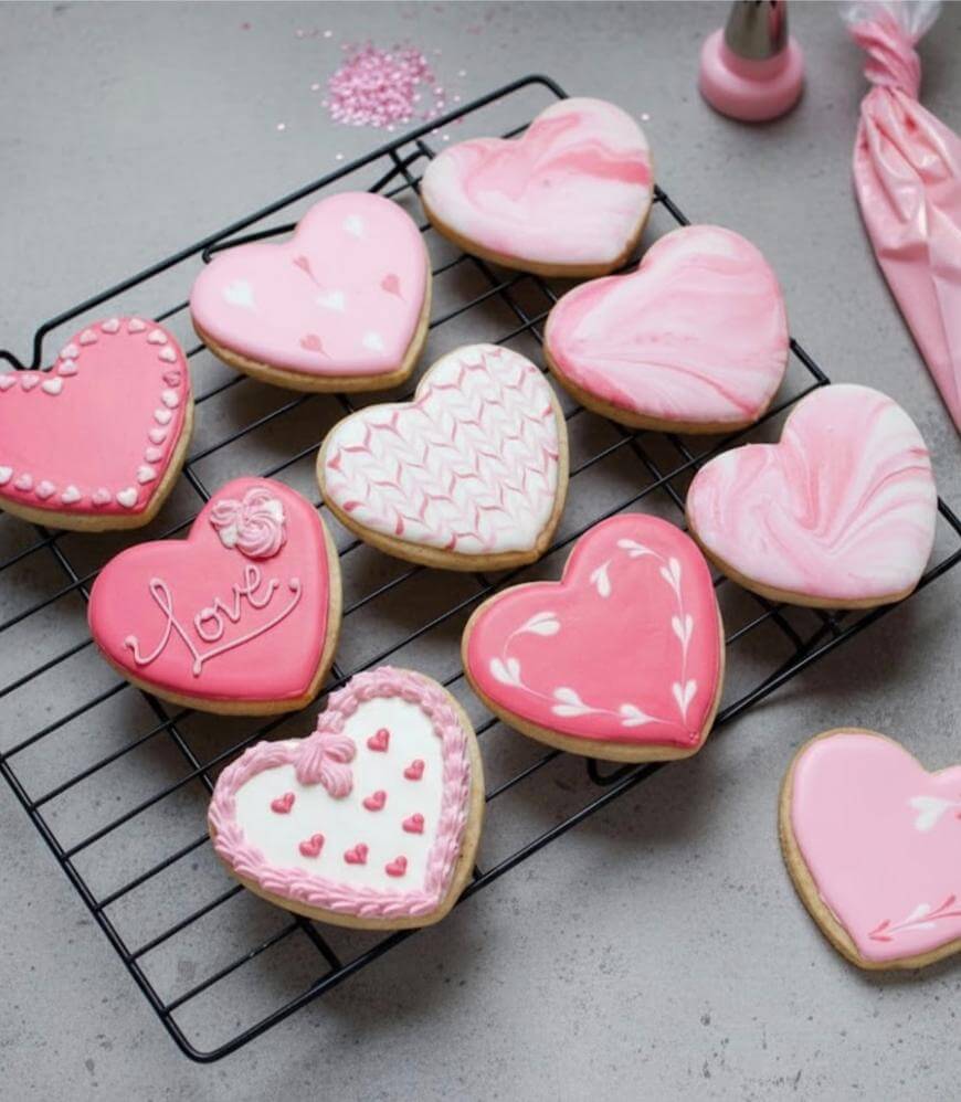 Heart-shaped sugar cookies decorated with pink and white icing sit on a black cooling rack. Each cookie has a unique design, featuring patterns, swirls, and the word Love. A piping bag and sprinkles are nearby.