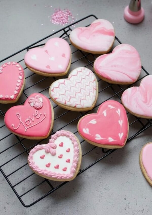 Heart-shaped cookies decorated with pink and white icing in various patterns are arranged on a cooling rack. The cookies have designs like hearts, swirls, and the word Love. Pink sprinkles and icing are in the background.