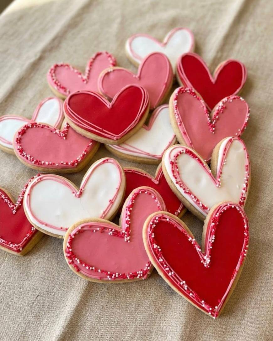 A pile of heart-shaped cookies decorated with red, pink, and white icing, some with sprinkles along the edges, arranged on a beige fabric surface.