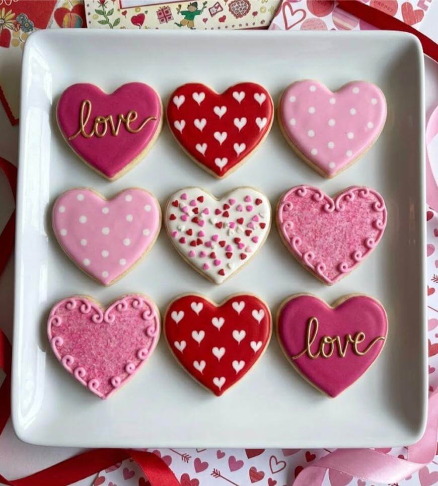A square white plate holds nine heart-shaped cookies, decorated in pink, red, and white frosting with patterns of hearts, dots, swirls, and the word “love” written on two cookies.