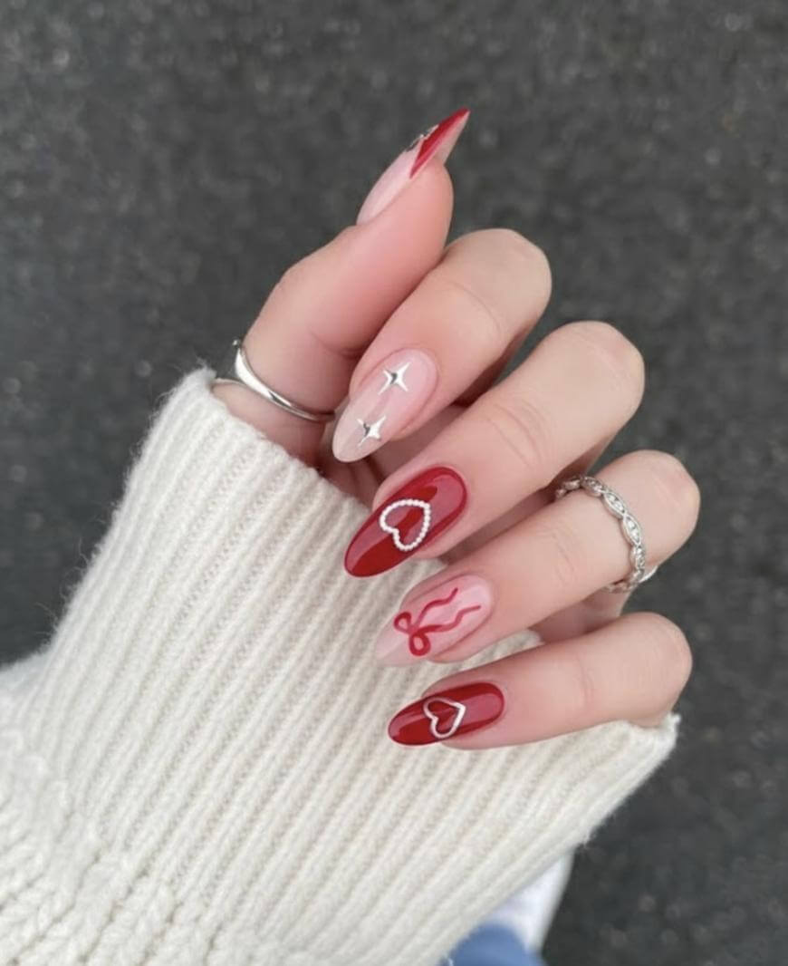 A hand with long manicured nails featuring red and pink nail art, including hearts and line designs. The person is wearing silver rings and a cream-colored knit sweater, with a gray textured background.