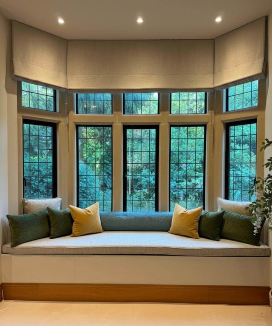 A cozy window seat with green and yellow cushions is set beneath large, multi-pane windows overlooking lush greenery. The area is softly lit by ceiling lights and framed with neutral Roman shades.