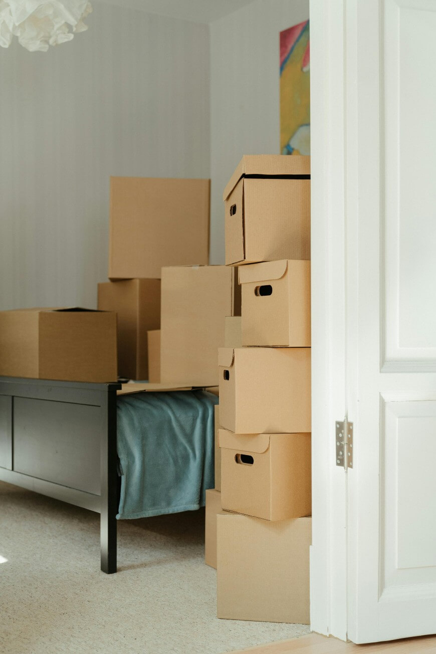 A bright room with a black table and several stacked cardboard moving boxes, some with lids and handles. Light filters in through an open white door, and a blue blanket covers part of the table.