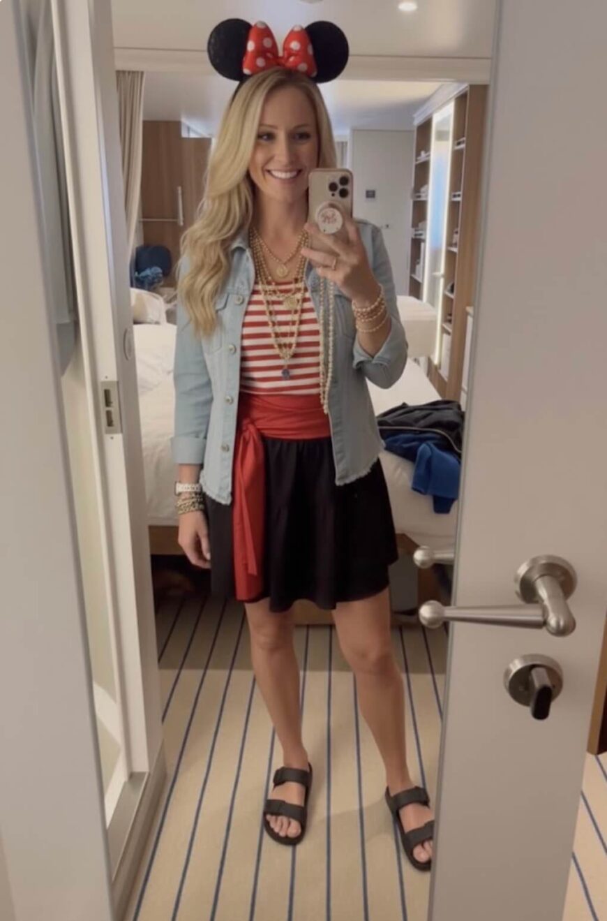 A woman stands in a bedroom taking a mirror selfie, wearing a Minnie Mouse ear headband, a striped shirt, a denim jacket, a red sash, a black skirt, and sandals. She is smiling and holding her phone.