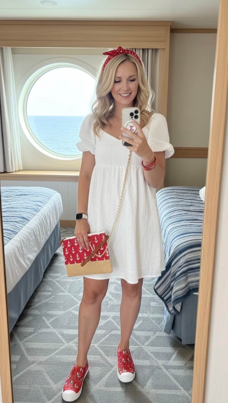 A woman in a white dress, red headband, red sneakers, and holding a red anchor-print purse takes a mirror selfie in a cabin with a round window showing the ocean. The cabin has two beds with blue-striped covers.