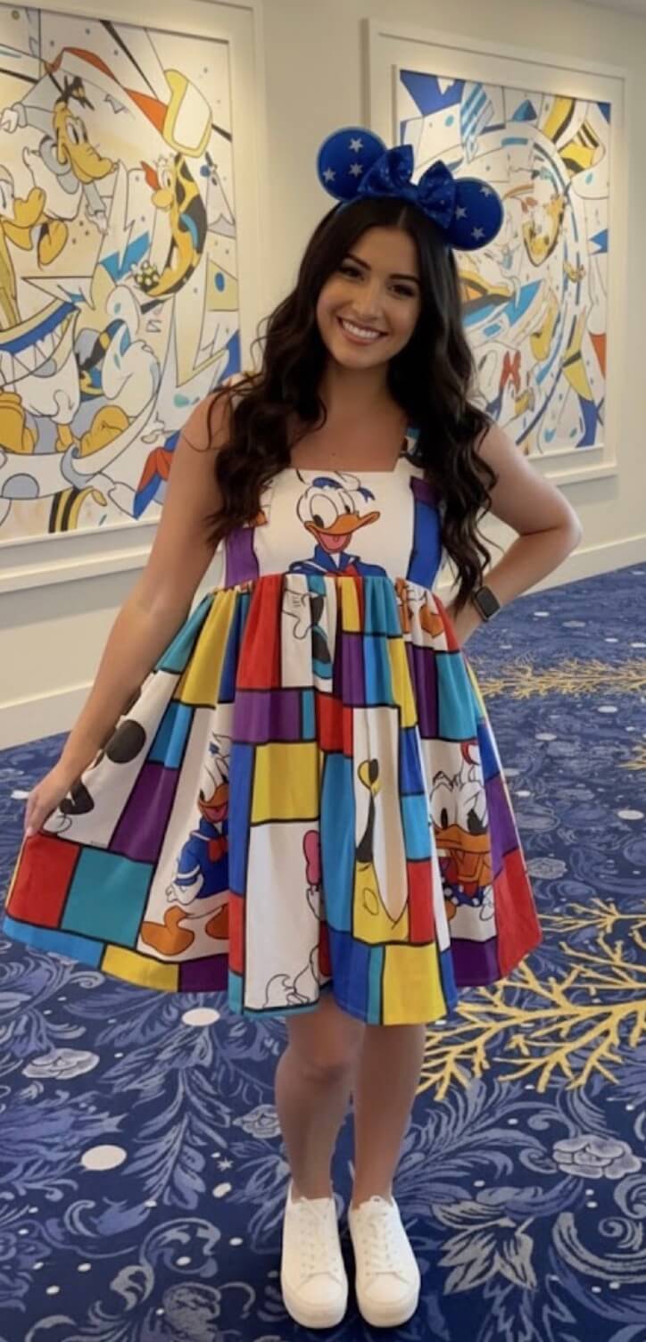 A woman smiles while wearing a colorful dress and blue Mickey Mouse ears. Her dress features a patchwork of cartoon images, including Donald Duck. She stands on a patterned blue carpet with Disney character art on the wall behind her.