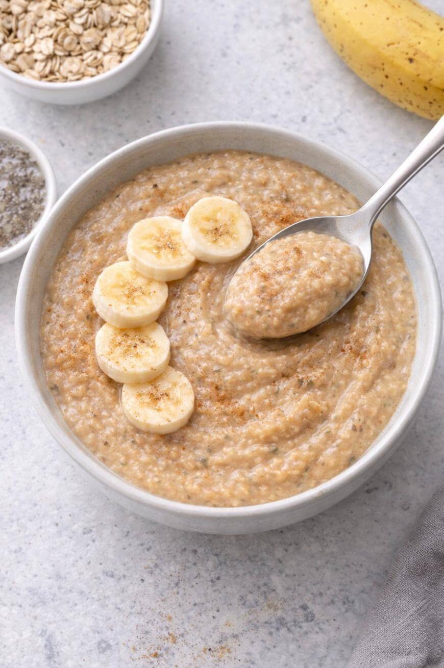 A bowl of creamy oatmeal topped with banana slices and a sprinkle of cinnamon, with a spoon scooping some out. Nearby are a banana, a bowl of oats, and a small dish of seeds on a light surface.