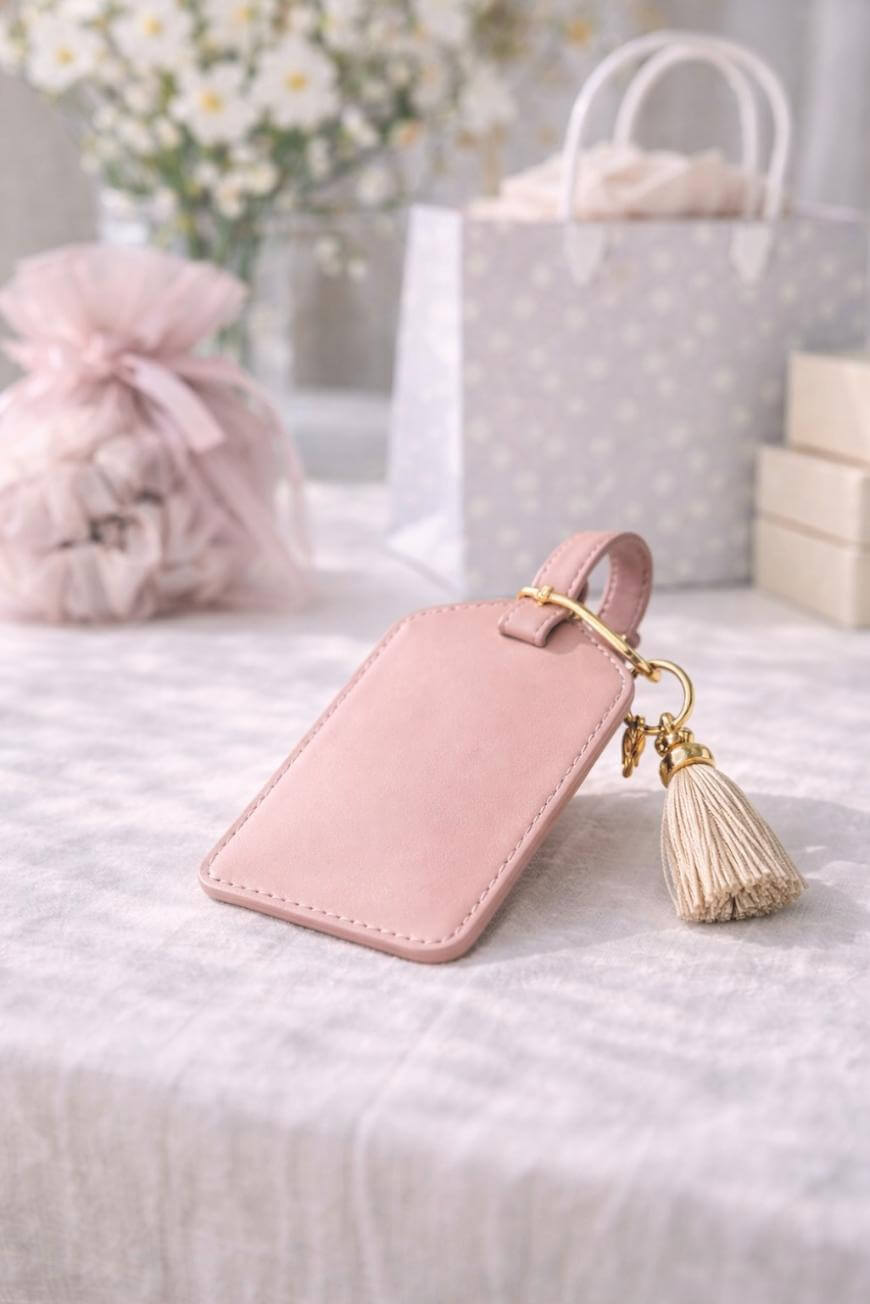 A soft pink leather luggage tag with a gold ring and a beige tassel rests on a light, textured tablecloth. In the background are pastel gift bags, boxes, and blurred white flowers.