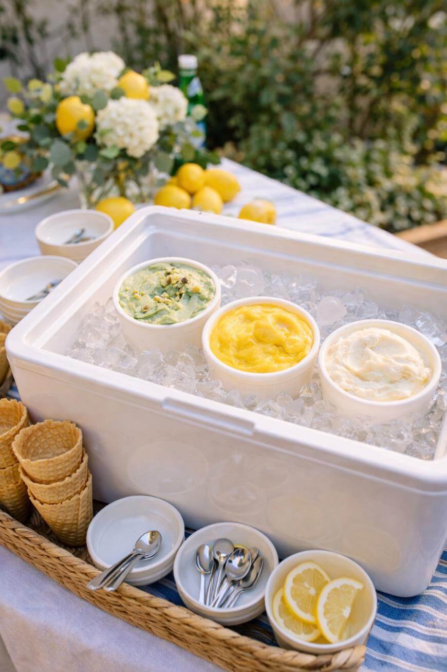 Three containers of colorful ice cream sit in a cooler of ice, surrounded by lemon slices, waffle cones, spoons, and bowls on a table decorated with fresh lemons and flowers.
