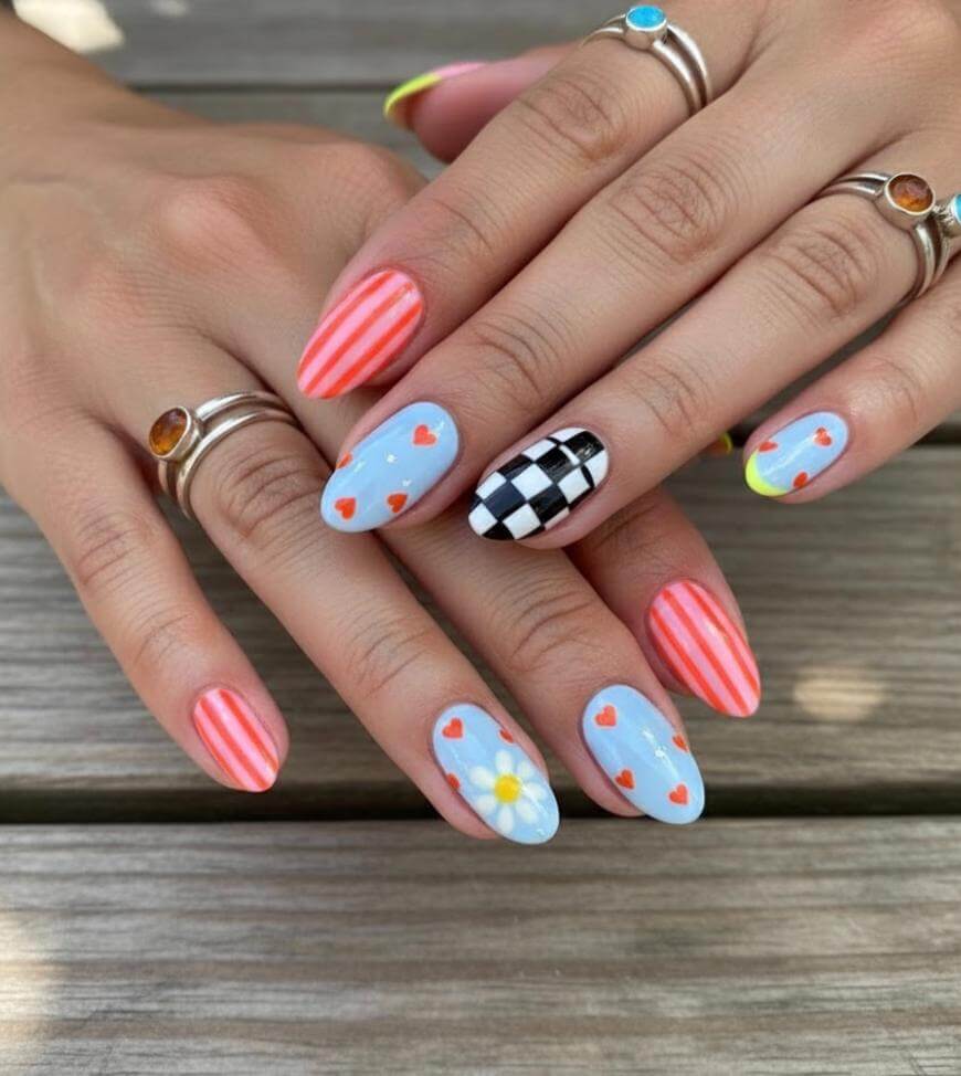 Hands with manicured nails featuring colorful designs: red stripes, blue with red hearts, a daisy flower, black-and-white checkerboard, and a yellow crescent. The person wears several silver rings. Nails are oval-shaped.