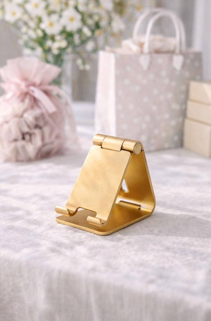 A gold metal phone stand sits on a light-colored tablecloth with blurred gift bags, boxes, and flowers in the background.