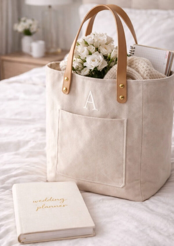 A beige tote bag with tan leather handles, monogrammed with the letter A, sits on a bed. Inside are white flowers, a knitted blanket, and a notebook. A wedding planner book lies in front of the bag.