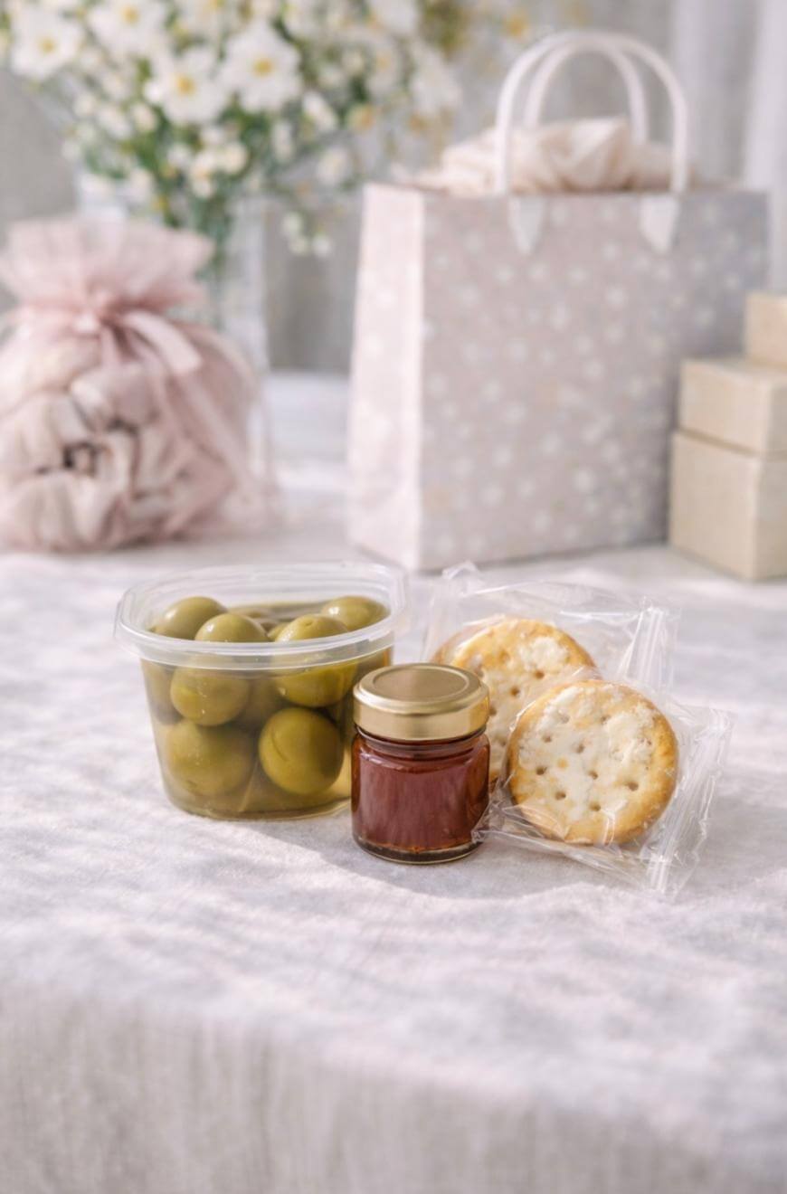 A small jar of jam, a container of green olives, and two round crackers in clear wrappers are arranged on a light tablecloth. Gift bags and boxes are in the blurred background.