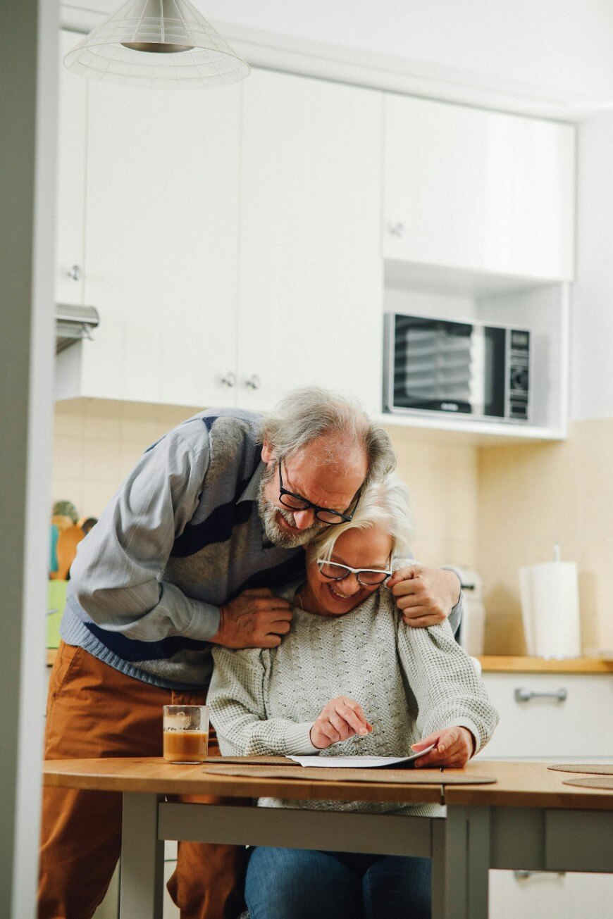 An elderly man hugs an elderly woman from behind as she sits at a kitchen table, smiling and looking at a tablet. They appear happy and relaxed in a bright, modern kitchen.