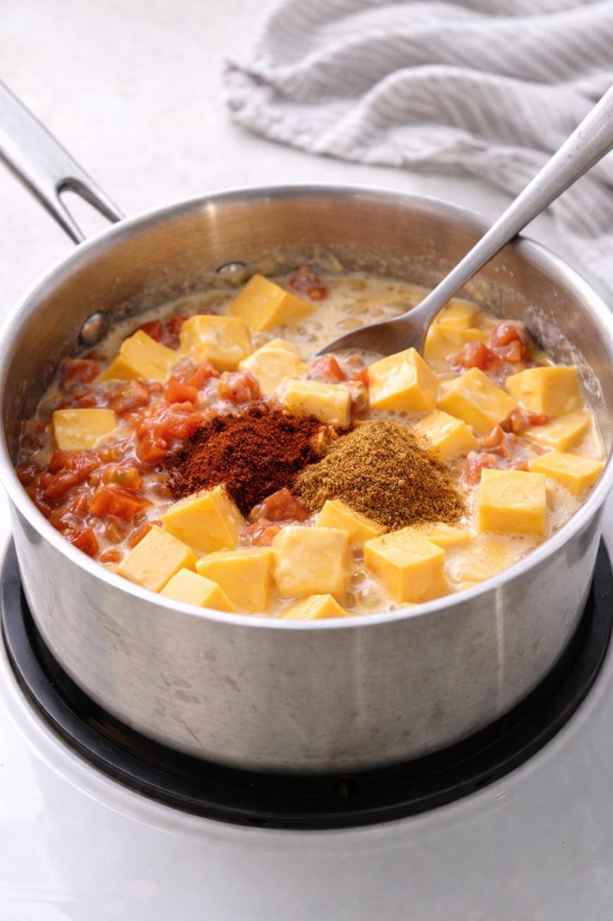 A stainless steel pot on a stovetop contains cubed cheese, diced tomatoes, and piles of spices being melted and mixed with a metal spoon. A striped cloth is in the background.
