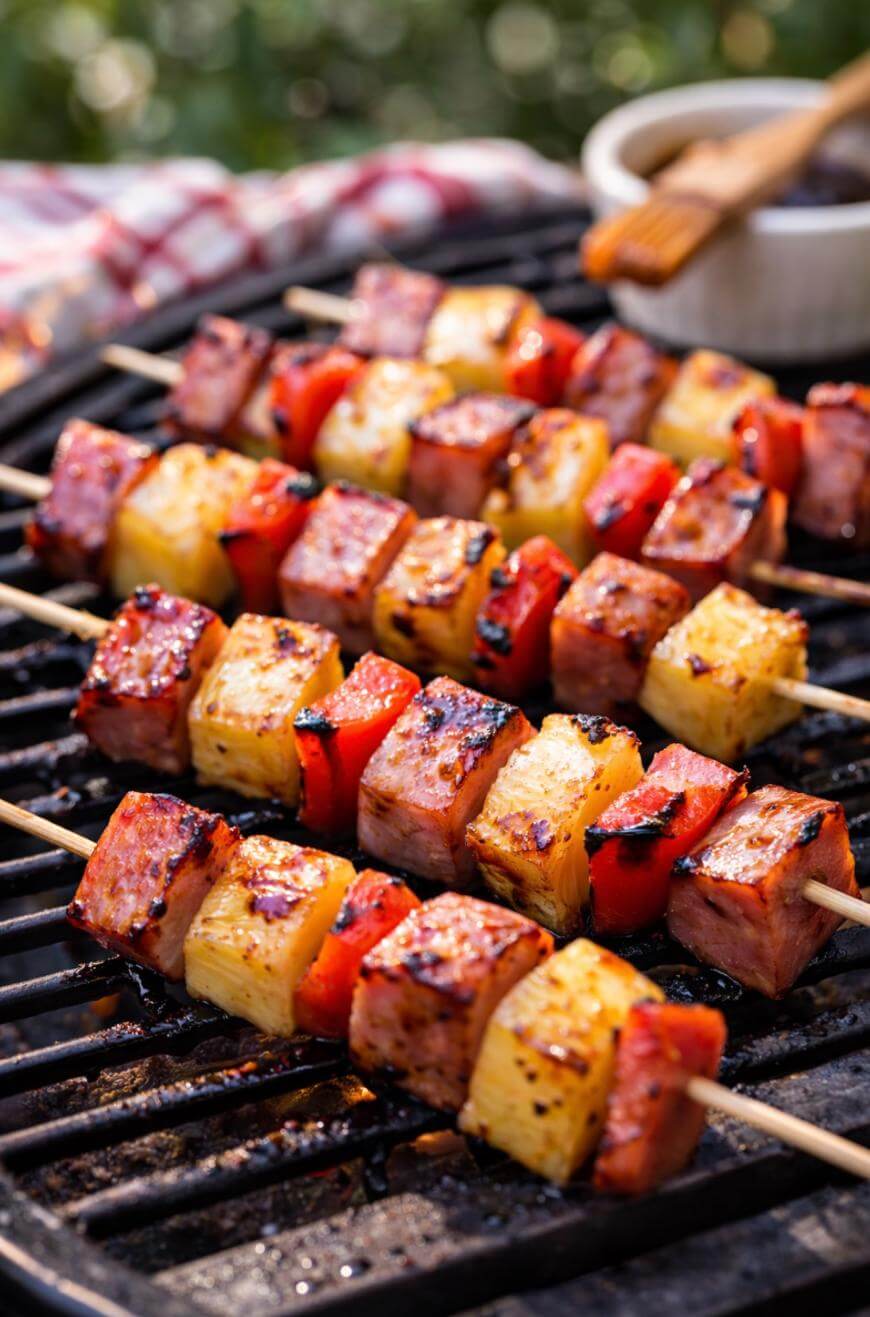 Grilled skewers with chunks of ham, pineapple, and red bell pepper cooking over a barbecue, with grill marks and a basting brush with sauce in the background.