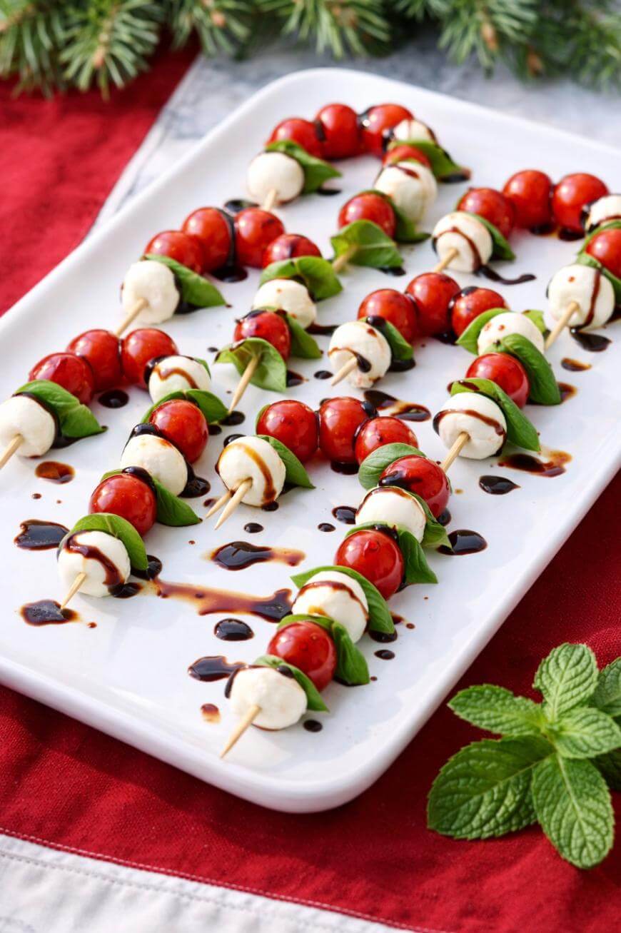 A white platter holds skewers of cherry tomatoes, mozzarella balls, and fresh basil leaves, drizzled with balsamic glaze. The platter is on a red and white cloth, with pine branches and mint as decoration.