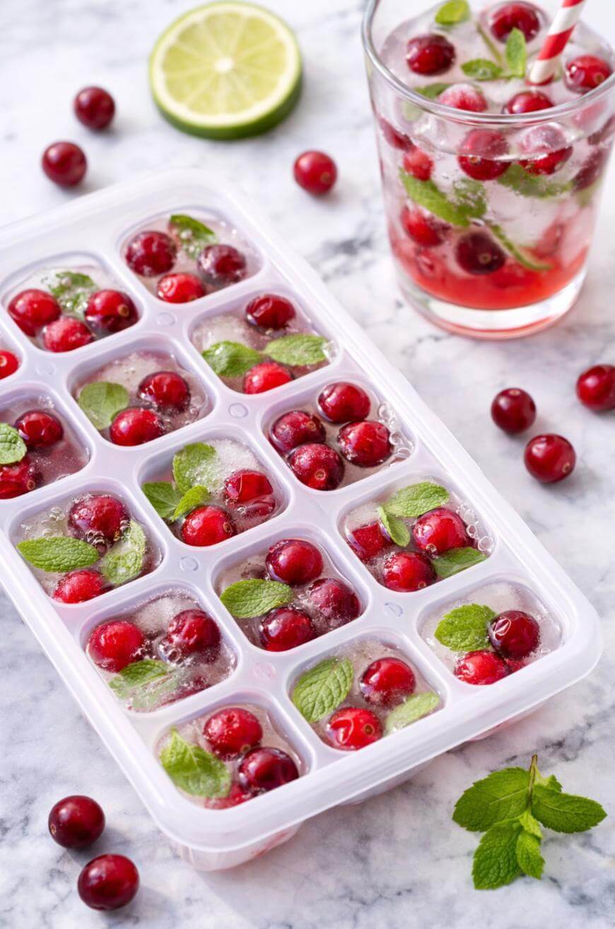 An ice cube tray filled with water, cranberries, and mint leaves; a glass of cranberry and mint drink with ice cubes and a striped straw; scattered cranberries, mint, and a lime slice on a marble surface.