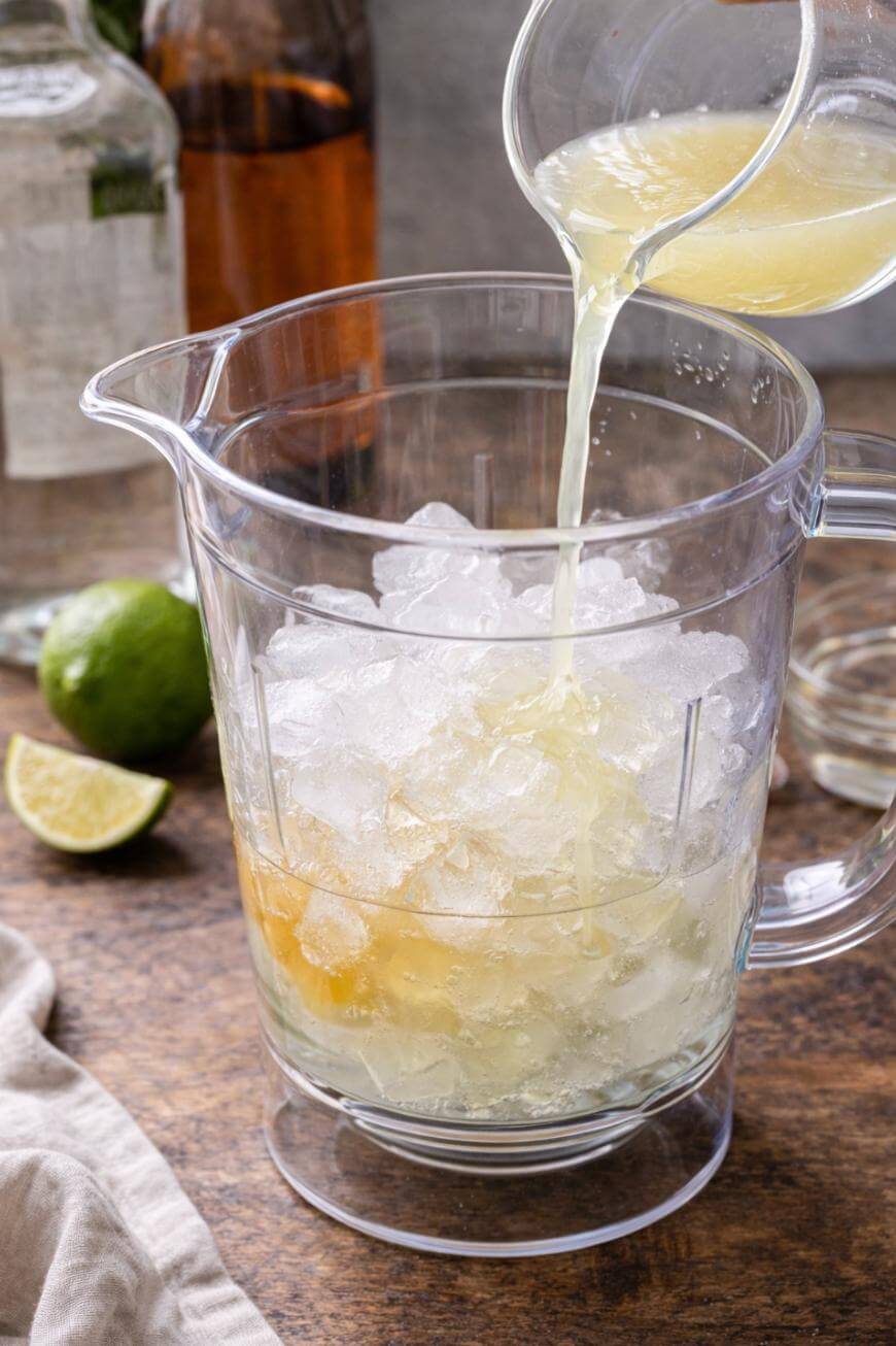 A clear pitcher filled with ice sits on a wooden surface as a hand pours yellow citrus juice into it. A sliced lime and bottles are visible in the background.