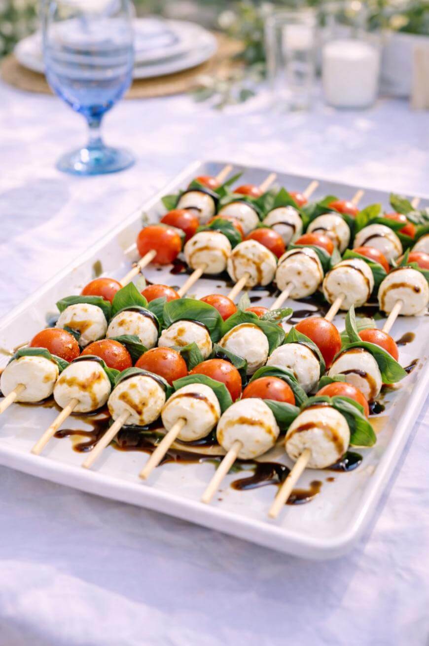 A platter of Caprese skewers with mozzarella balls, cherry tomatoes, and basil leaves, drizzled with balsamic glaze, is set on an outdoor table with a blue glass and plates in the background.