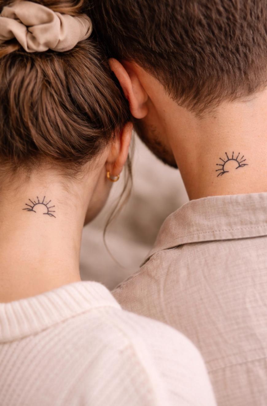 Two people with their backs turned, each showing a small matching tattoo of a half sun with rays on the nape of their necks. One has hair tied with a scrunchie; both wear light-colored clothing.