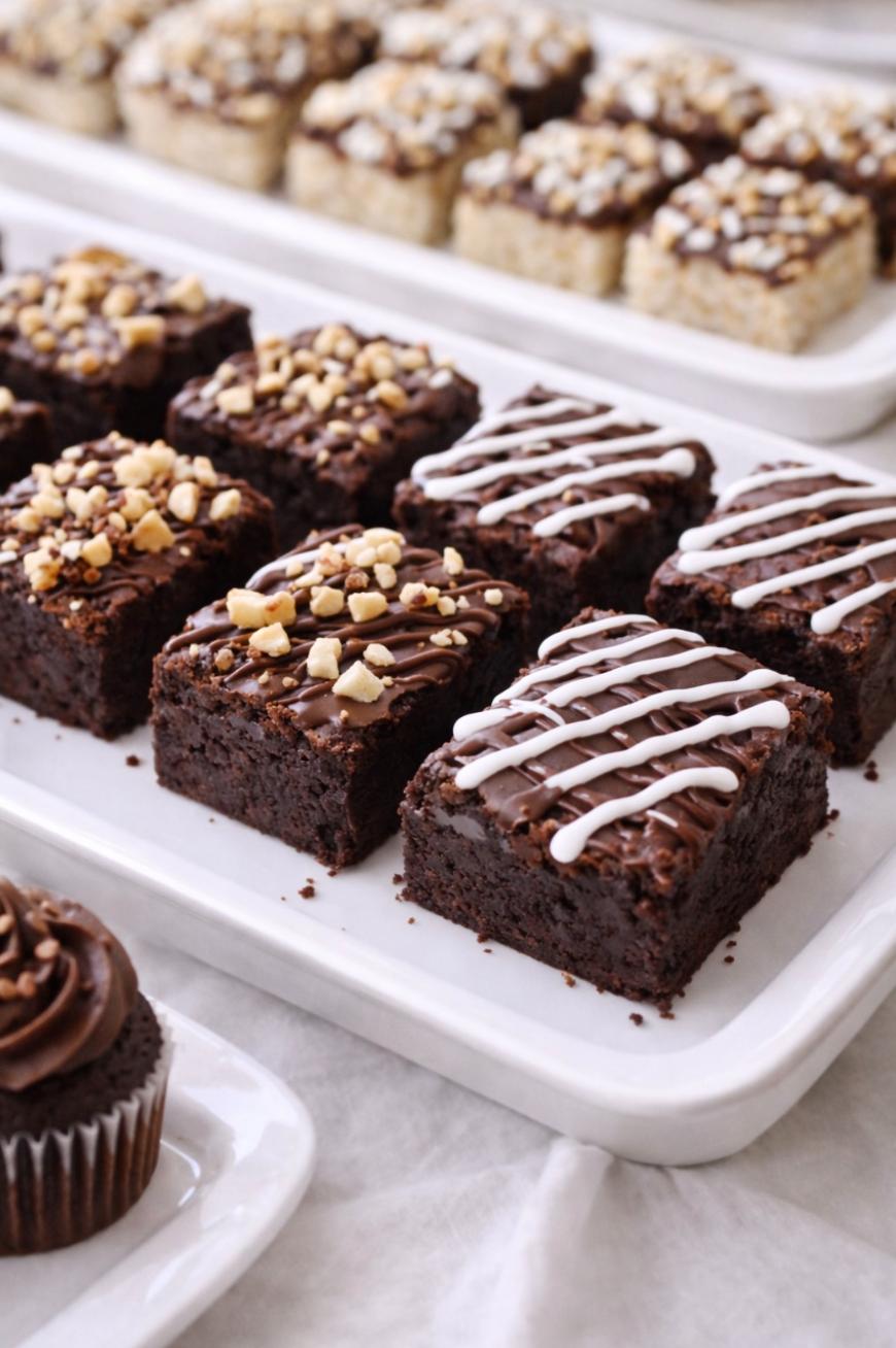 A tray of chocolate brownies, some topped with chopped nuts and others drizzled with white icing, with more assorted brownies and a chocolate cupcake in the background.