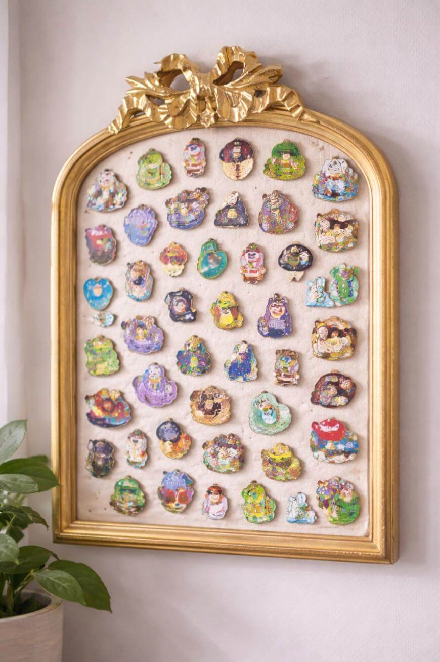 A gold-framed display case hangs on a wall, holding several rows of painted decorative pins or brooches, each featuring colorful cartoon-like characters. A green plant sits in the lower left corner.