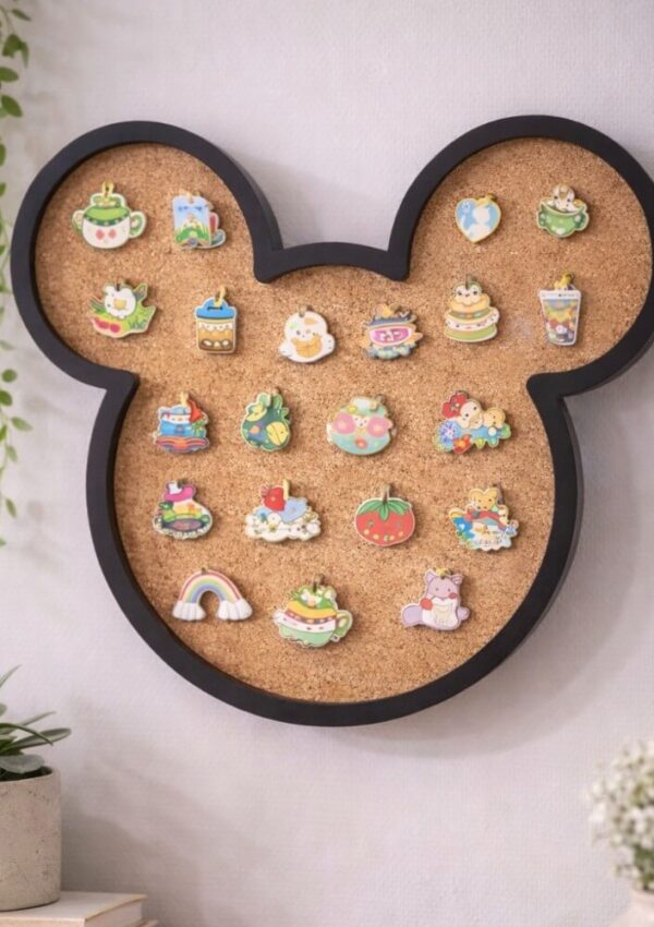 A corkboard shaped like Mickey Mouse’s head is mounted on a wall and decorated with colorful, cute pins featuring food, animals, and cheerful designs. Green potted plants sit on a white surface nearby.