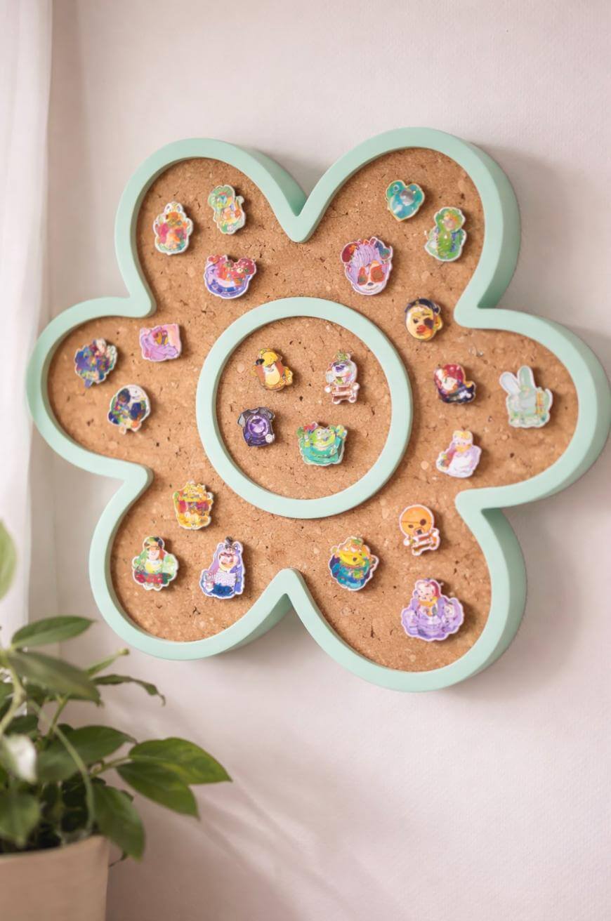 A flower-shaped corkboard with a mint green border displays colorful enamel pins, featuring various cartoon characters. A green potted plant sits nearby on the left.