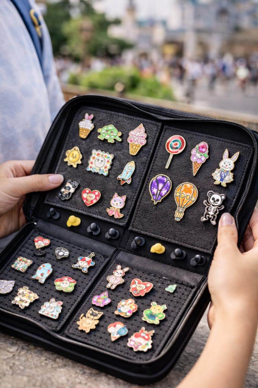 A person holds open a case displaying a colorful collection of enamel pins, including animals, ice cream cones, balloons, and various cute characters, set against an outdoor, blurred background.