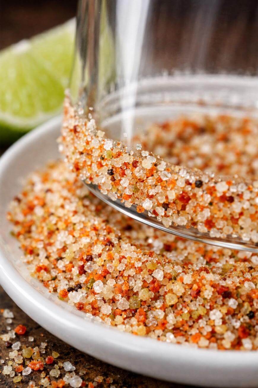 A glass rim being dipped into a plate of coarse, colorful seasoning salt, with a wedge of lime in the background.