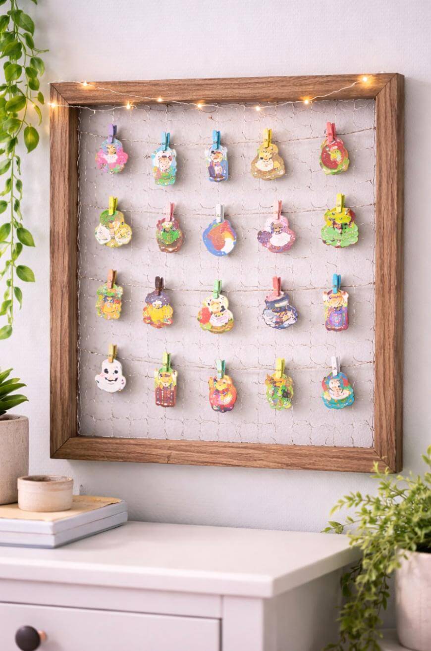 A wooden frame with wire holds small, colorful charms clipped to it in rows; the frame is decorated with string lights and hangs on a light wall above a shelf with plants and decor.