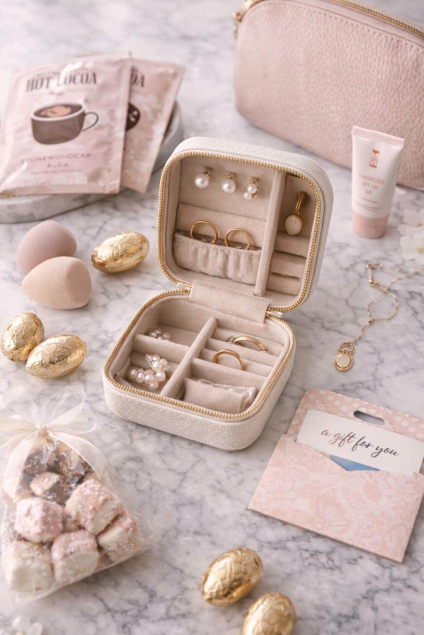 A beige jewelry box with rings, earrings, and a pearl necklace sits on a marble surface, surrounded by gold-wrapped chocolates, beauty products, a gift tag, pink marshmallows, and a blush-colored pouch.
